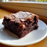 A close-up of a rich, fudgy square of chocolate brownies dusted lightly with powdered sugar on a white plate.