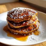 A close-up of a stack of three moist Gingerbread pancakes drizzled with syrup and dusted with powdered sugar.