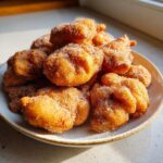 A plate piled high with freshly fried, golden brown Buñuelos coated generously in granulated sugar.