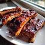 Four pieces of tender, glazed BBQ ribs with a rich, dark sauce piled on a white plate.