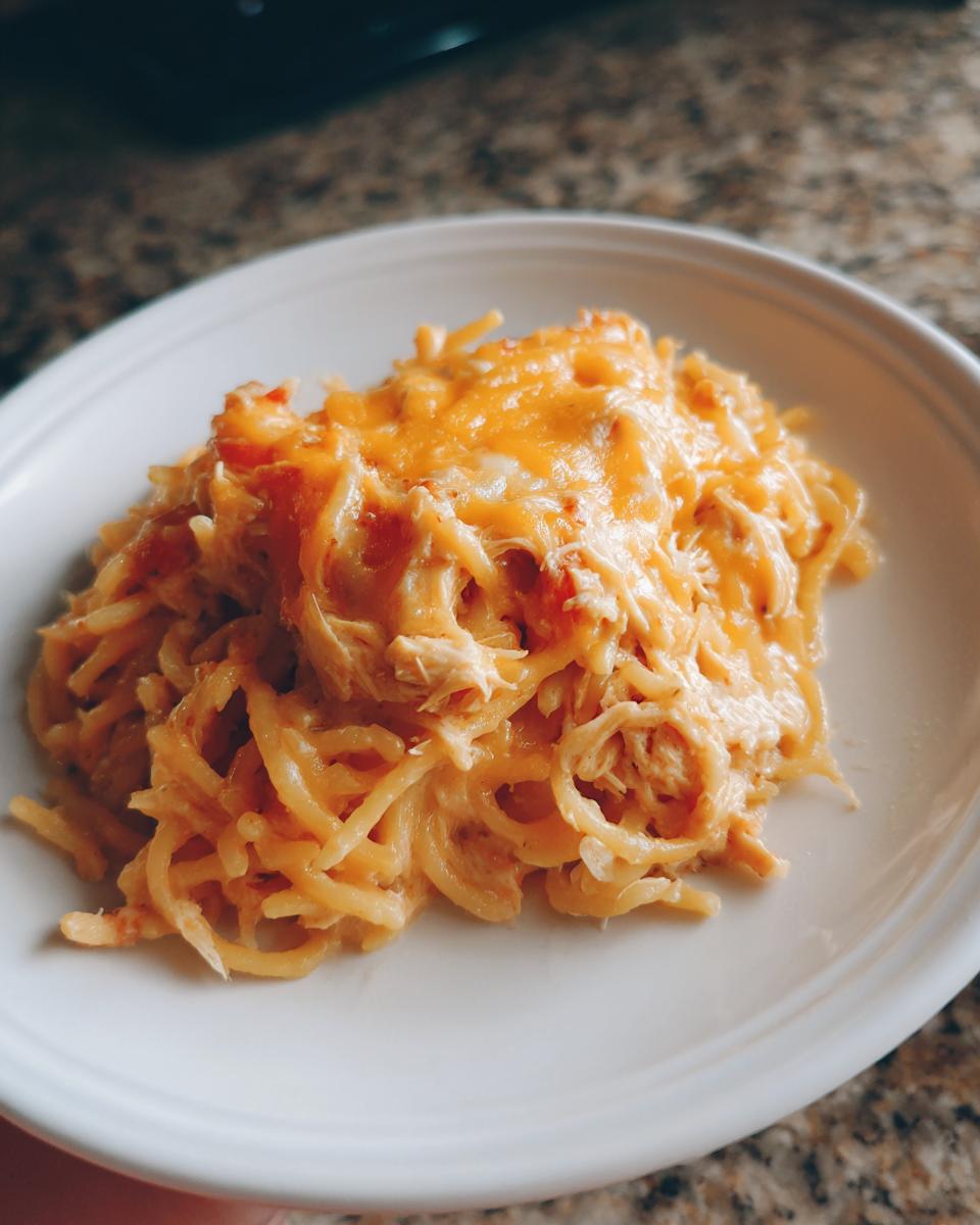 A close-up of a portion of Monterey chicken spaghetti topped with melted orange cheddar cheese on a white plate.