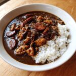 A close-up of a bowl of rich, dark Gumbo featuring chicken pieces and sliced andouille sausage served alongside fluffy white rice.