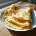 A stack of thin, golden-brown crepes, folded into quarters, resting on a white plate.