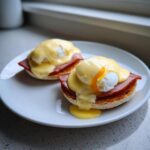Two servings of classic eggs benedict featuring poached eggs, ham, and rich hollandaise sauce on toasted English muffins.