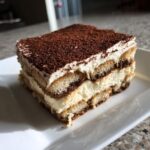 A close-up of a layered slice of Tiramisu, featuring coffee-soaked ladyfingers and creamy mascarpone, dusted with cocoa powder.