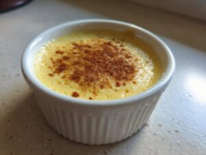 Close-up of a creamy yellow egg custard dessert dusted with cinnamon in a white fluted ramekin.