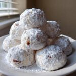 A stack of freshly baked Snowball cookies heavily coated in powdered sugar, resting on a white plate.