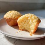 Two moist vanilla cupcakes on a white plate, one of which has a bite taken out, showing the fluffy interior.