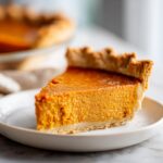 A close-up of a slice of delicious vegan pumpkin pie on a white plate, showcasing its rich orange filling and flaky crust.