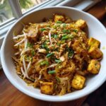 A close-up of a white bowl filled with delicious vegetarian Pad Thai, featuring rice noodles, crispy tofu cubes, bean sprouts, peanuts, and green onions.