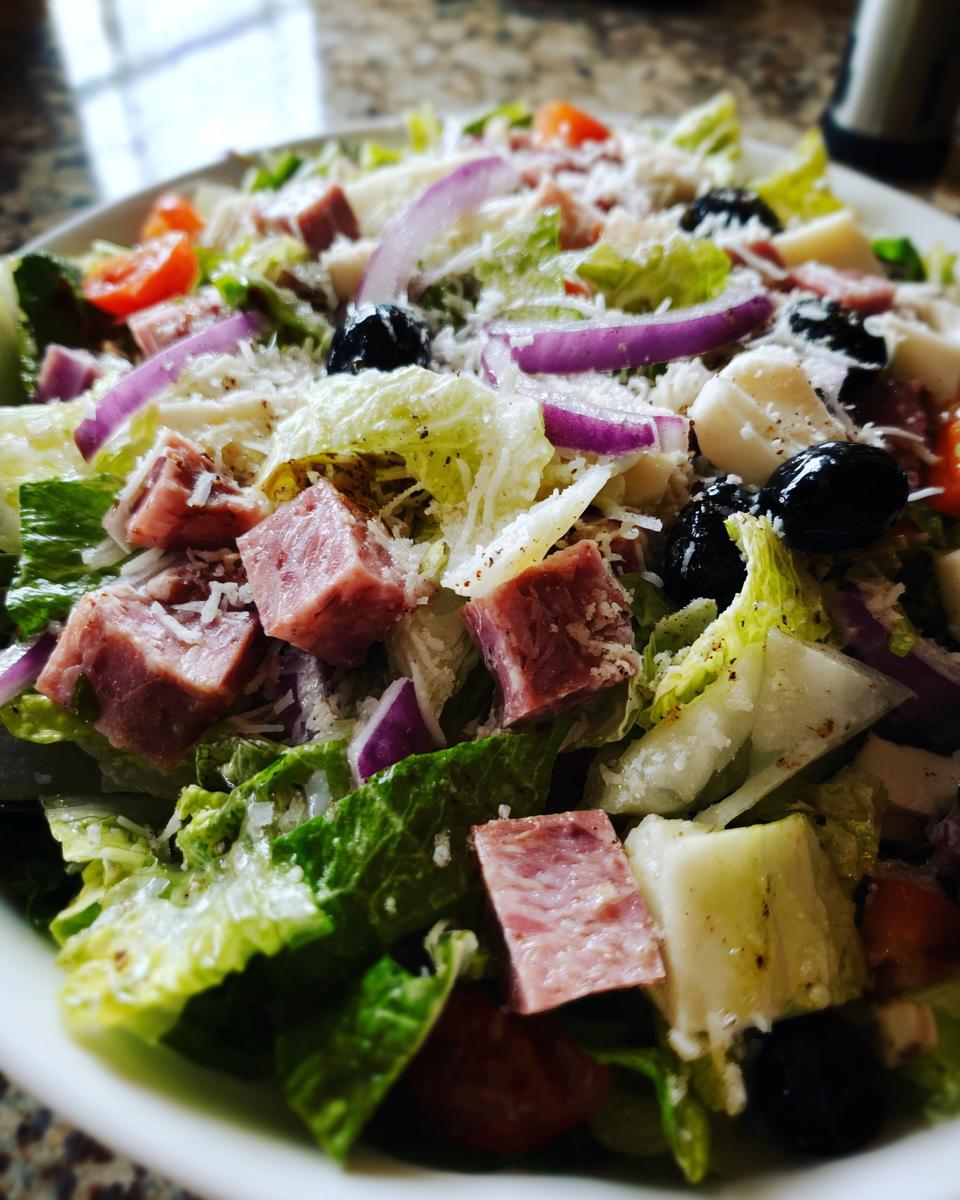 Close-up of a vibrant grinder salad featuring lettuce, cubed deli meats, red onion, black olives, and grated cheese.