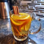 A clear mug filled with warm apple cider, garnished with orange slices, cinnamon sticks, and whole cloves.