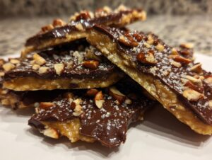 Close-up of broken pieces of homemade toffee recipe, topped with dark chocolate, chopped nuts, and sea salt flakes.