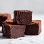 Three rich, dark chocolate fudge squares stacked on a light marble surface, showcasing the dense texture of the fudge.