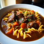Close-up of a rich, dark bowl of beef noodle soup with tender chunks of beef, carrots, and egg noodles.