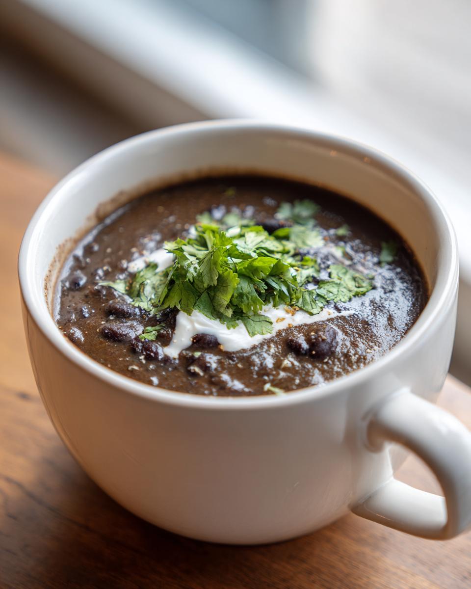 Close-up of a creamy black bean soup in a white mug, garnished with sour cream and fresh cilantro.