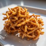A close-up of two golden-brown, glossy haystack cookies made from crunchy chow mein noodles coated in caramel.