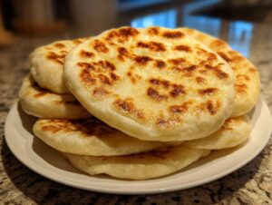 A stack of freshly cooked, puffy pita bread rounds with beautiful golden-brown charred spots on a white plate.