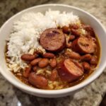 A close-up of a white bowl filled with savory red beans and rice topped with thick slices of smoked sausage.