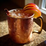 A condensation-covered copper mug filled with an apple cider moscow mule, garnished with apple slices and a cinnamon stick.