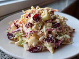 Close-up of a serving of creamy apple cranberry coleslaw featuring shredded cabbage, carrots, apples, and dried cranberries.