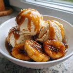 Three scoops of vanilla ice cream topped with caramel sauce next to caramelized bananas in a bowl of Bananas Foster.