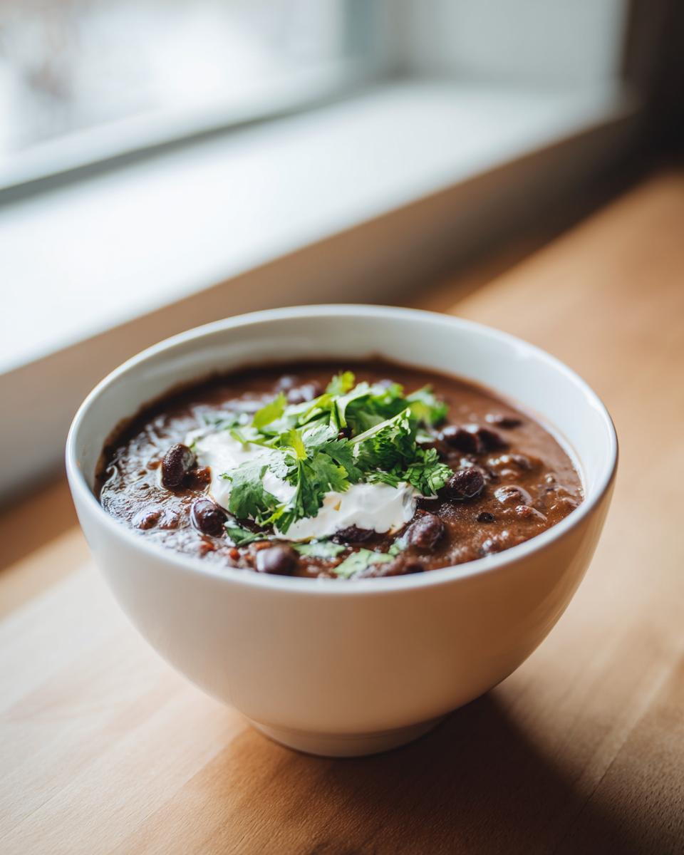 A white bowl filled with thick, dark black bean soup, topped with sour cream and fresh cilantro.