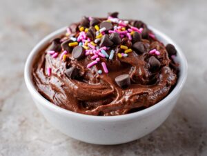 Close-up of rich, thick brownie batter dip in a white bowl, topped with chocolate chips and colorful sprinkles.