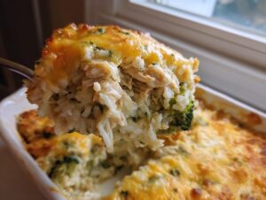A spoonful of creamy chicken broccoli rice casserole topped with melted, golden cheese being lifted from the baking dish.