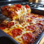 A serving of hot pepperoni pizza dip is lifted, showing long, gooey cheese strings stretching back to the pan.
