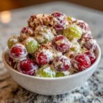 A white bowl filled with creamy Chicken Salad Chick Grape Salad, featuring red and green grapes coated in dressing and topped with pecans.