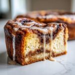 A close-up of a moist slice of cinnamon roll cheesecake with rich cinnamon swirl and dripping vanilla icing.