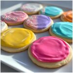 Close-up of sugar cookies topped with vibrant, glossy cookie icing in pink, yellow, purple, and blue.