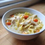 A close-up of a white bowl filled with creamy chicken soup, featuring egg noodles, shredded chicken, and bright orange carrots.