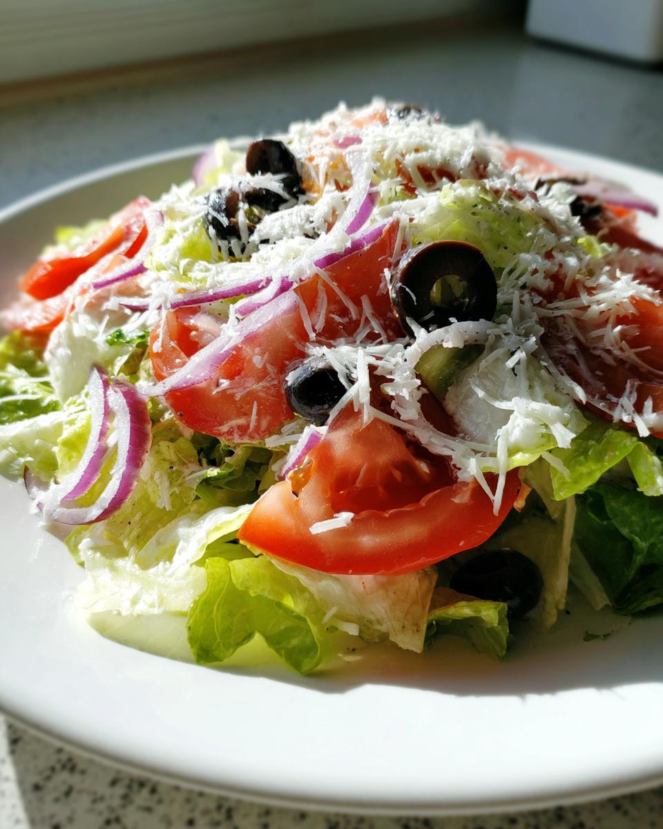A close-up of a fresh copycat Olive Garden salad featuring crisp lettuce, tomato slices, red onion, black olives, and grated Parmesan cheese.