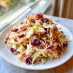 A mound of creamy apple cranberry coleslaw, featuring shredded cabbage and bright red dried cranberries, served on a white plate.