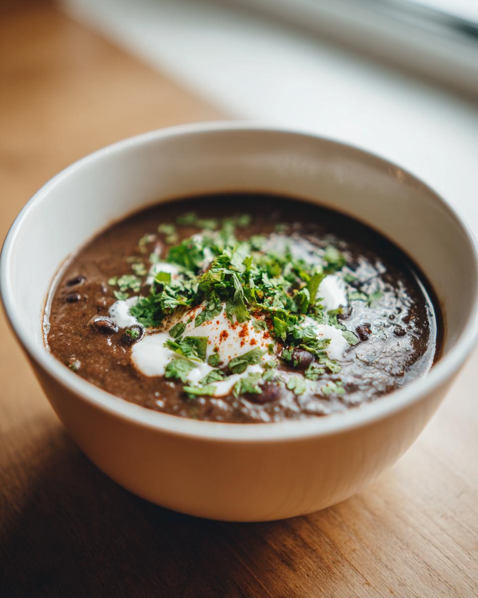 A close-up of a bowl of creamy black bean soup topped with sour cream, fresh cilantro, and paprika.