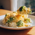 A spoonful of creamy broccoli rice lifted from a plate, showing rich cheese sauce and bright green broccoli florets.