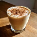 A glass of creamy coquito recipe topped generously with ground cinnamon, sitting on a wooden table.