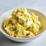 A close-up of a creamy, chunky egg salad recipe served in a white bowl, featuring chopped celery and bacon.