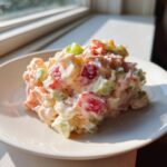 A mound of creamy seafood salad featuring colorful diced ingredients served on a white plate near a sunlit window.