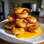 A stack of golden brown, crispy coconut shrimp served on a white plate with a pool of bright orange dipping sauce.