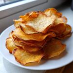 A tall stack of golden brown, crispy homemade sweet potato chips seasoned with pepper on a white plate.