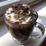 A clear glass mug filled with rich crockpot hot chocolate, topped high with whipped cream and chocolate shavings.