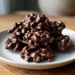 A close-up stack of glossy, dark chocolate crockpot peanut clusters resting on a light gray plate.