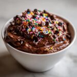 A close-up of rich, edible brownie batter dip topped generously with chocolate chips and colorful rainbow sprinkles.