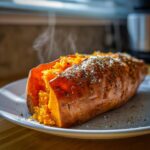 A fluffy, steaming baked sweet potatoes split open and seasoned with salt and herbs on a white plate.