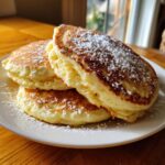 A stack of three fluffy greek yogurt pancakes dusted generously with white powdered sugar on a white plate.