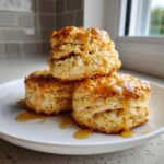 Three fluffy sweet potato biscuits stacked and drizzled generously with golden honey on a white plate.