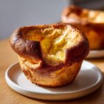 A single, tall, and fluffy Yorkshire pudding with a deep golden-brown crust sitting on a speckled white plate.